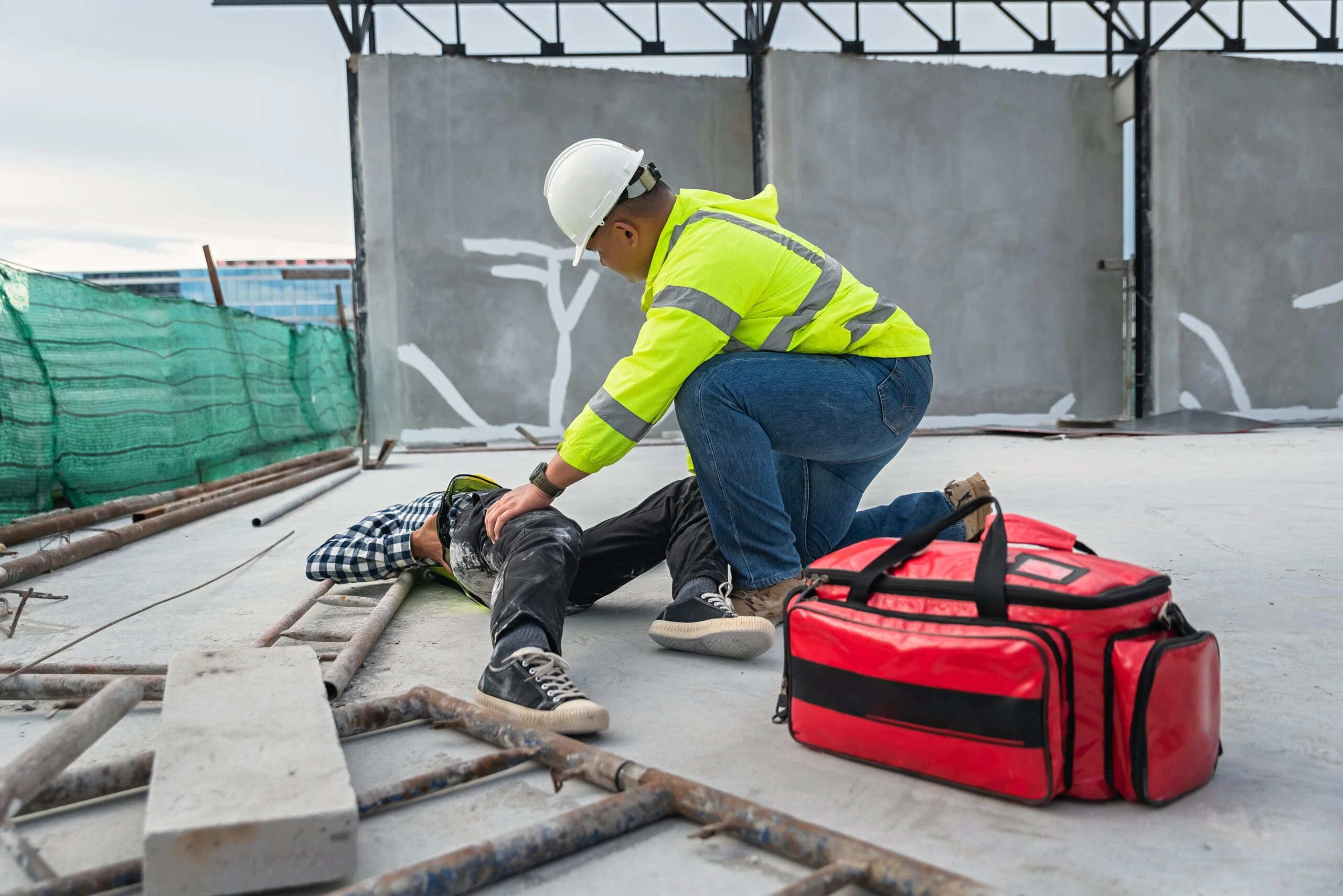 man in hi vis jacket and hard hat treating an employee on the floor with a red first aid bag in the forefront.