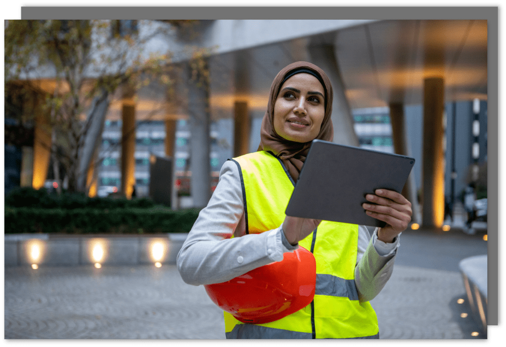 A women standing with a yellow hi vis and a red hard hat under her arm holding and using a tablet.