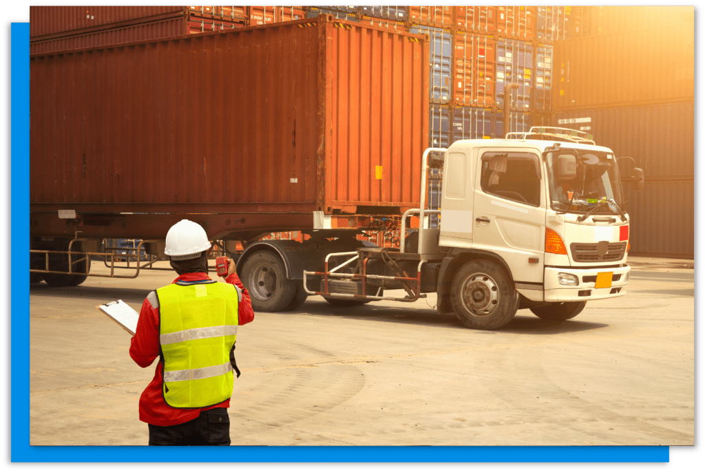 A worker standing in front of a lorry in a pallet yard wearing a yellow hi vis vest and a white hard hat.