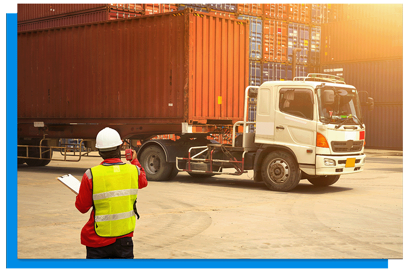 A worker standing in front of a lorry in a pallet yard wearing a yellow hi vis vest and a white hard hat.