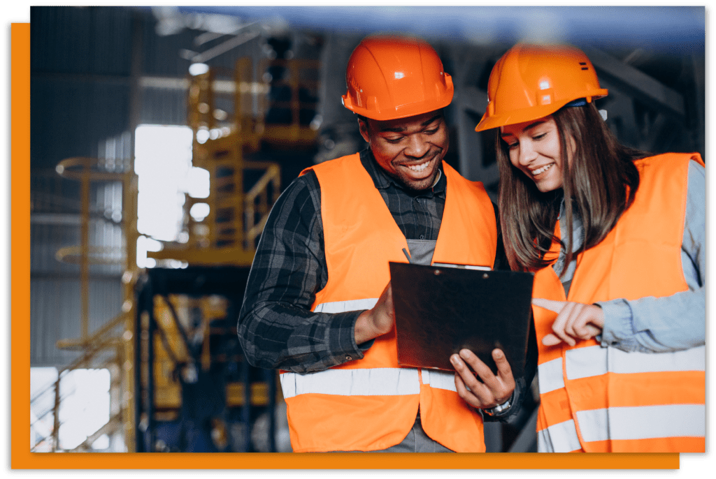 two workers in orange hard hats and hi vis vests looking at a tablet in a warehouse.