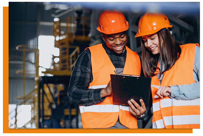 two workers in orange hard hats and hi vis vests looking at a tablet in a warehouse.