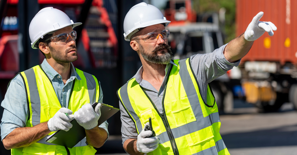 Two workers on a construction site in hard hats and hi vis vests. One is pointing into the distance.