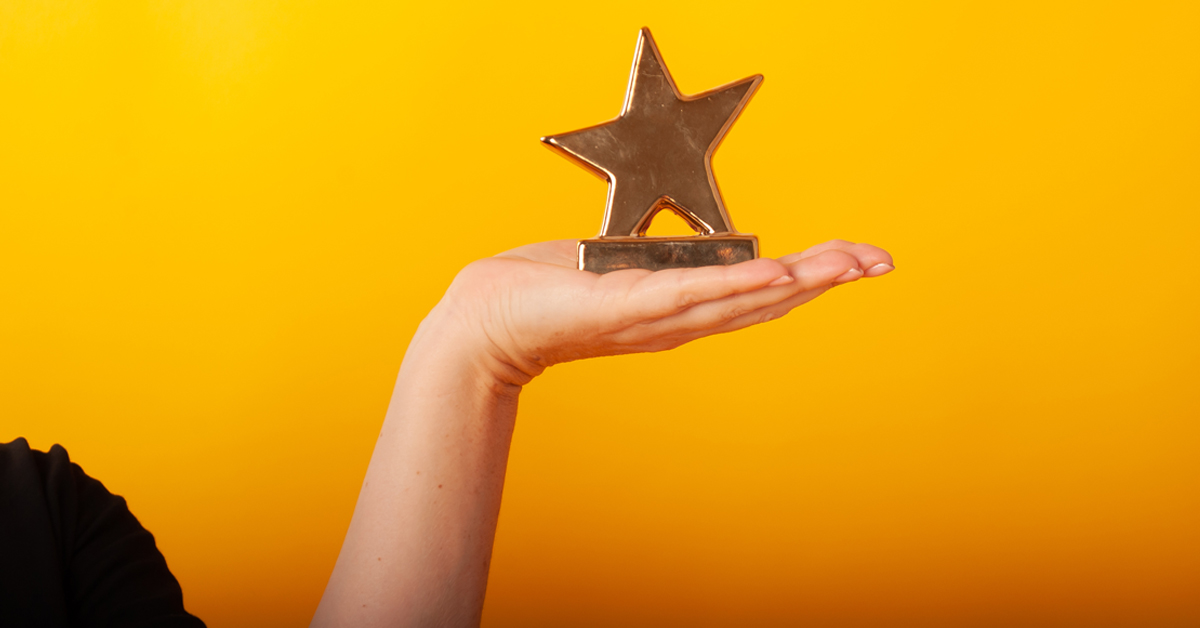 A hand holding a gold star award in front of a bright yellow backdrop.