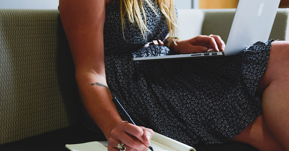 Female worker sitting on a sofa at a laptop writing on a notepad.