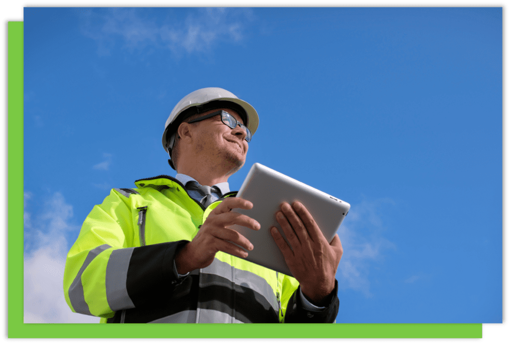 Image taken from below looking up at a man in a hardhat and hi vis jacket holding a tablet looking into the distance.