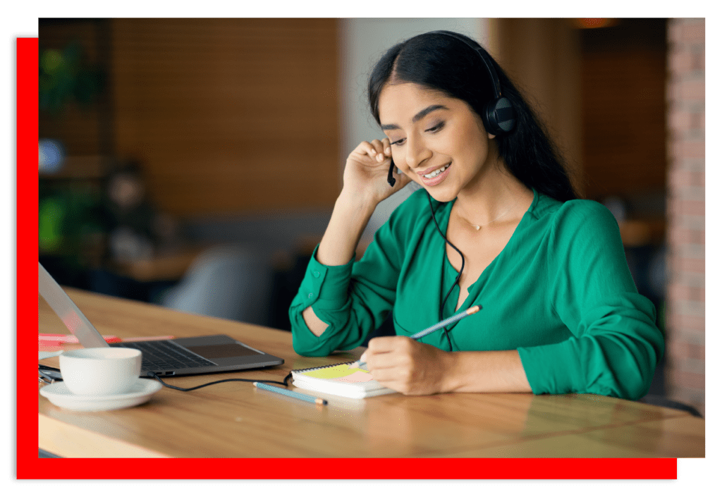 Lady wearing a green silk blouse with a headset on talking whilst taking notes with a laptop and a coffee cup on the table.