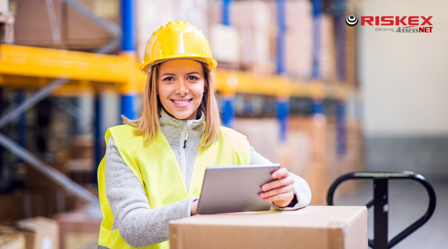A worker in a bright vest and hard hat using a tablet while handling a box in a warehouse.