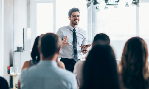 Man presenting to a group of people in business attire carrying a tablet.