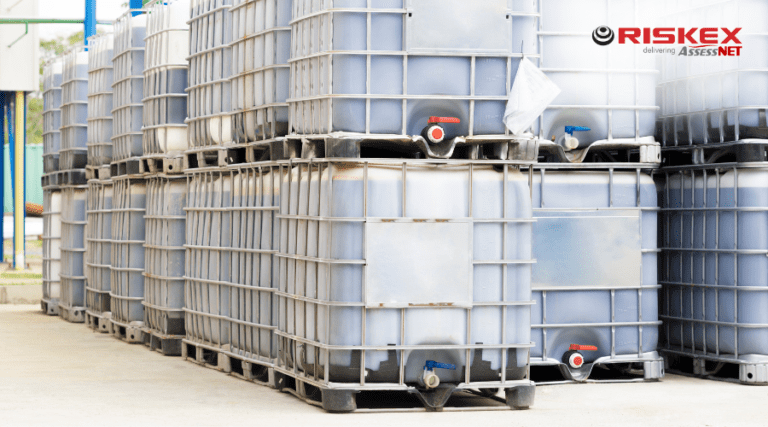 Stacked industrial IBC containers filled with liquid chemicals, secured in metal cages with visible valves, in a warehouse setting.